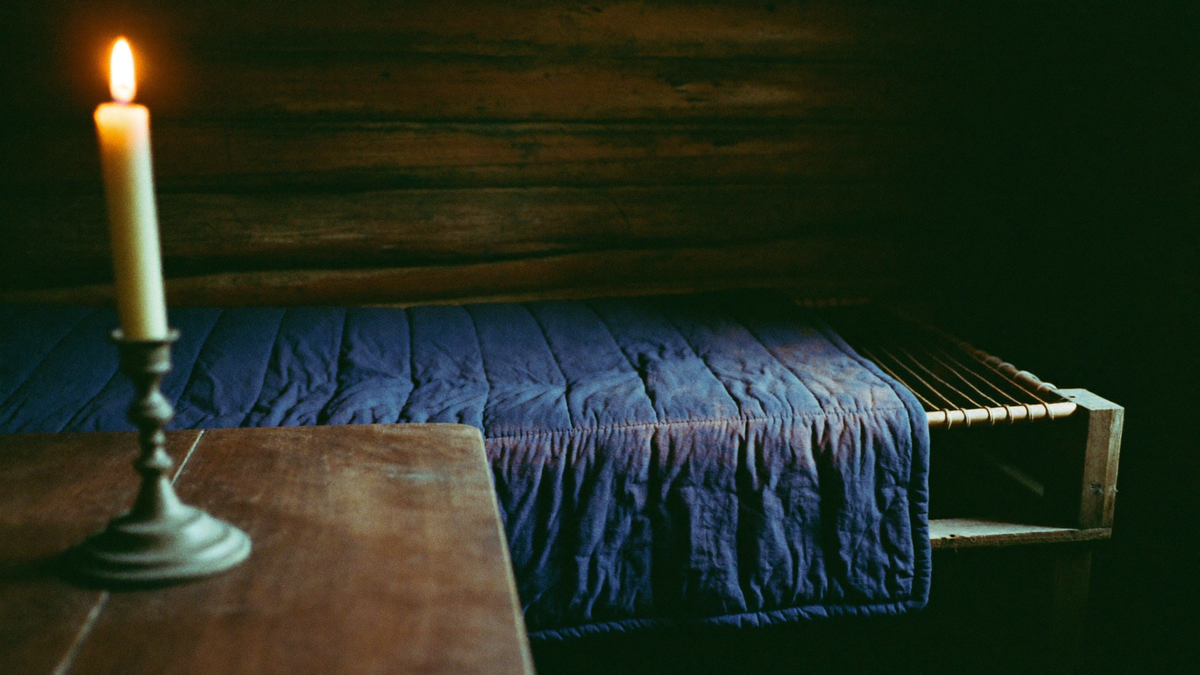 Candle burning on a wooden table beside a quilted rope bed inside a dark early 1800s log cabin