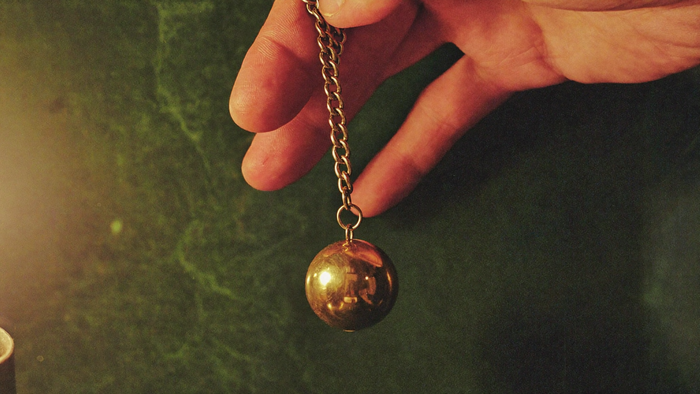 A hand holds a brass pendulum on a chain above a dark green surface, caught mid-swing
