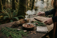 Hands fanning cedar smoke with a feather over a bowl of cedar chips and fresh cedar fronds in a forest