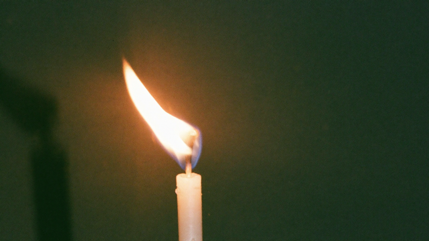 A single candle flame leaning and dancing to one side against a dark green background