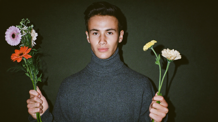 A young man in a grey turtleneck holding two small bunches of flowers against a dark green wall