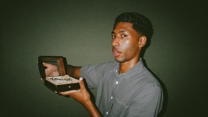 A young man in a grey shirt holding an open secondhand wooden jewelry box against a dark green wall