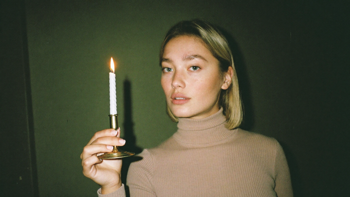 A woman in a plain turtleneck holding a lit taper candle in a brass holder against a dark green wall