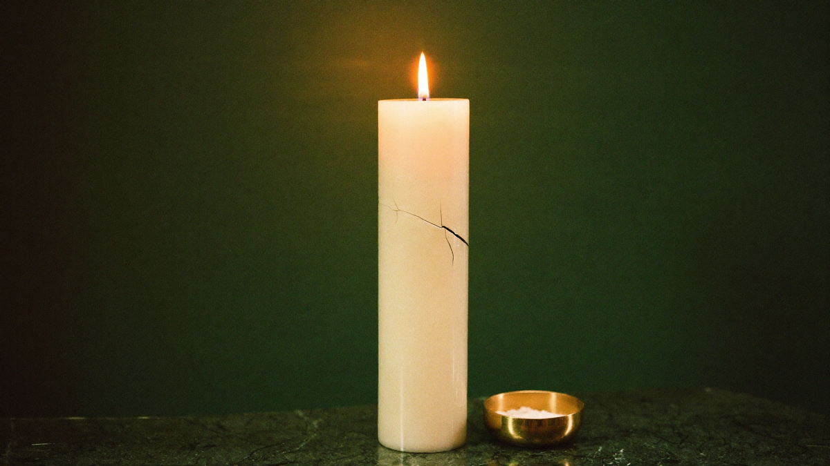 A tall vigil candle with a hairline crack in its side, burning on a dark green stone altar surface