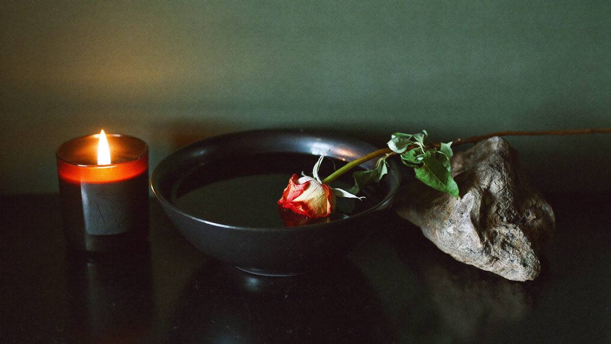 A lit candle, a dark bowl of still water and a wilting red rose on a black surface in dim green light