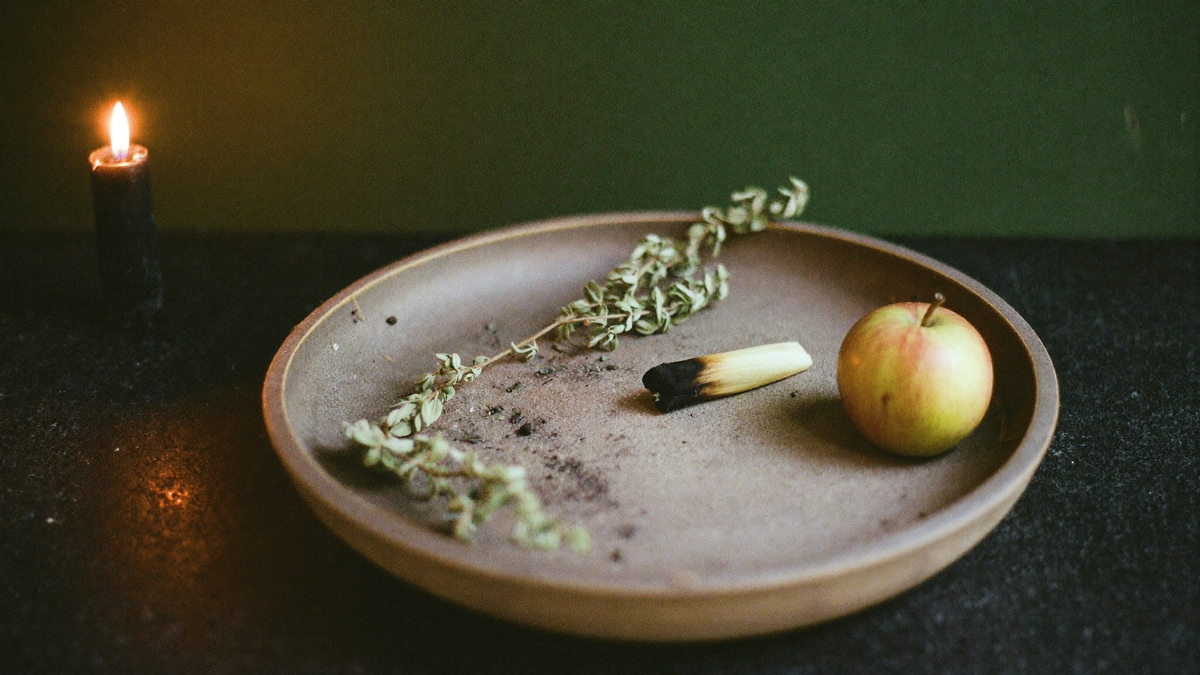 A ceramic bowl of ash holding dried herbs, a burned-out candle stub and an apple on dark stone, lit by one candle