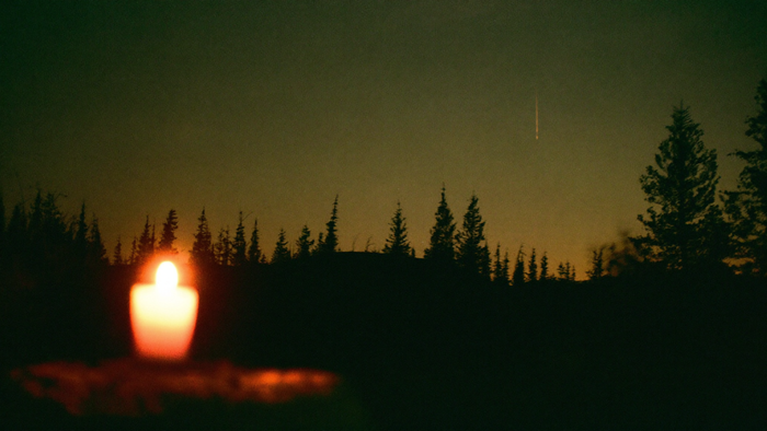 Single meteor streak in the dusk sky above a dark ridge of spruce trees, a lit candle glowing in the foreground