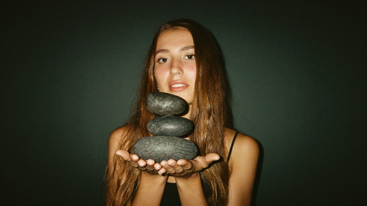 A young woman holding four stacked dark grey stones balanced in her open hands against a deep green wall