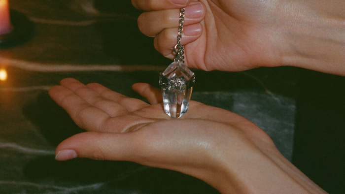 Close-up of a clear crystal pendulum on a chain held over an open palm in warm candlelight