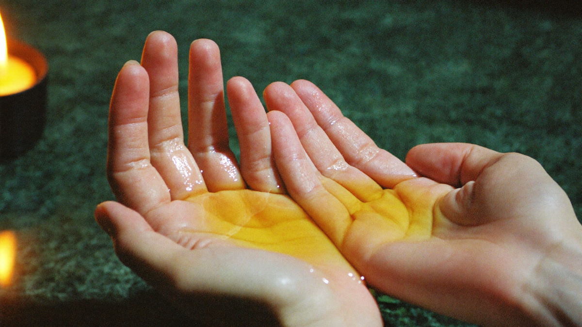 Two cupped hands holding a splash of pale golden cologne, lit by a candle against dark green stone