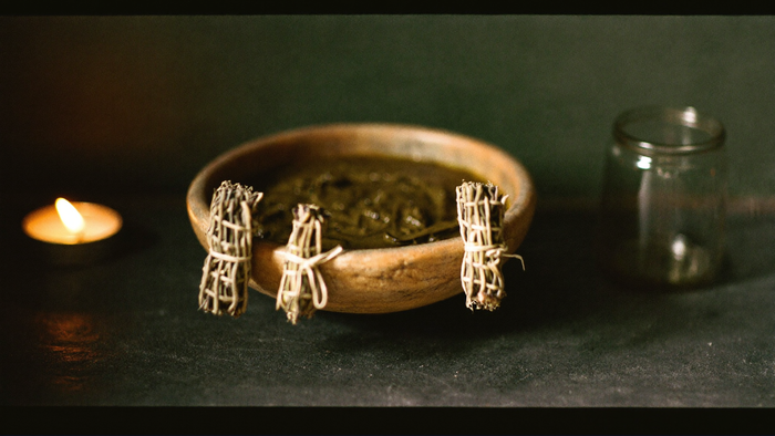 A wooden bowl of dark green herb paste with three tied herb bundles and a glass jar on dark stone