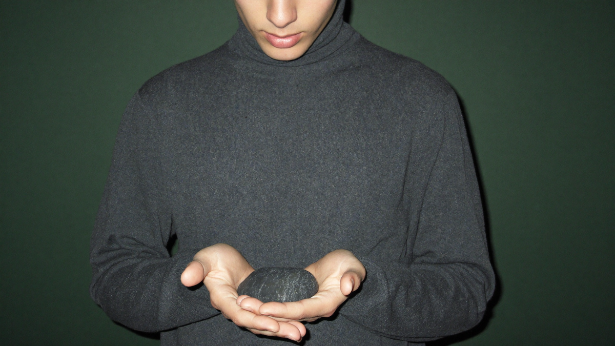 A young man in a plain grey turtleneck holding a single dark tumbled stone in cupped hands