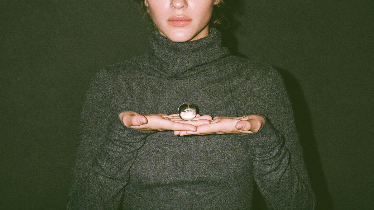 A young woman in a grey turtleneck holding a small clear crystal ball in both hands against a dark green wall