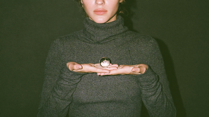 A young woman in a grey turtleneck holding a small clear crystal ball in both hands against a dark green wall