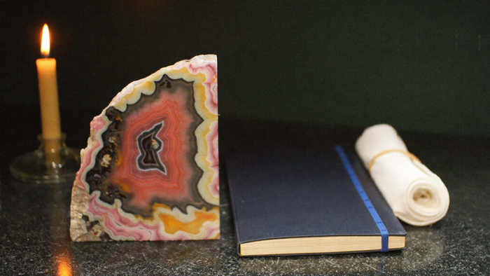 A pink banded stone beside a dark notebook and a folded cloth on a stone surface, lit by a single candle