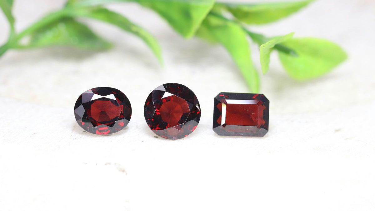 Three faceted deep red garnets, oval, round and emerald cut, on a pale stone surface with soft green leaves behind