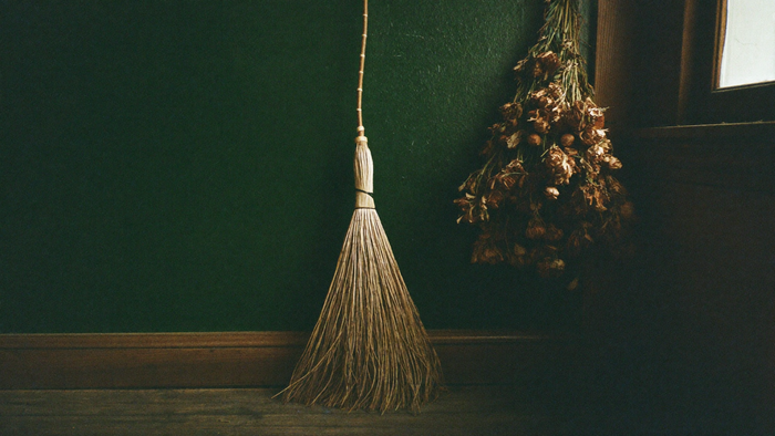 Handmade twig besom leaning against a dark green wall beside a bundle of dried herbs in soft morning light