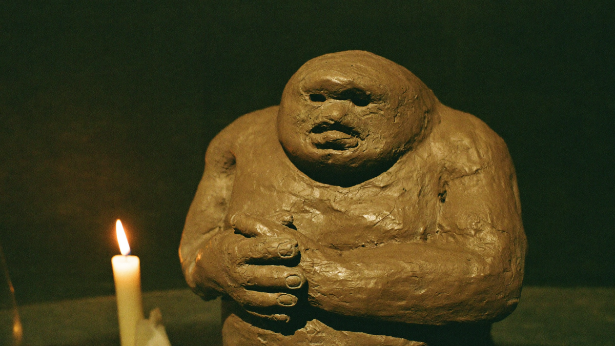 Roughly shaped clay golem figure with a heavy head, hollow eyes and folded hands beside a lit candle