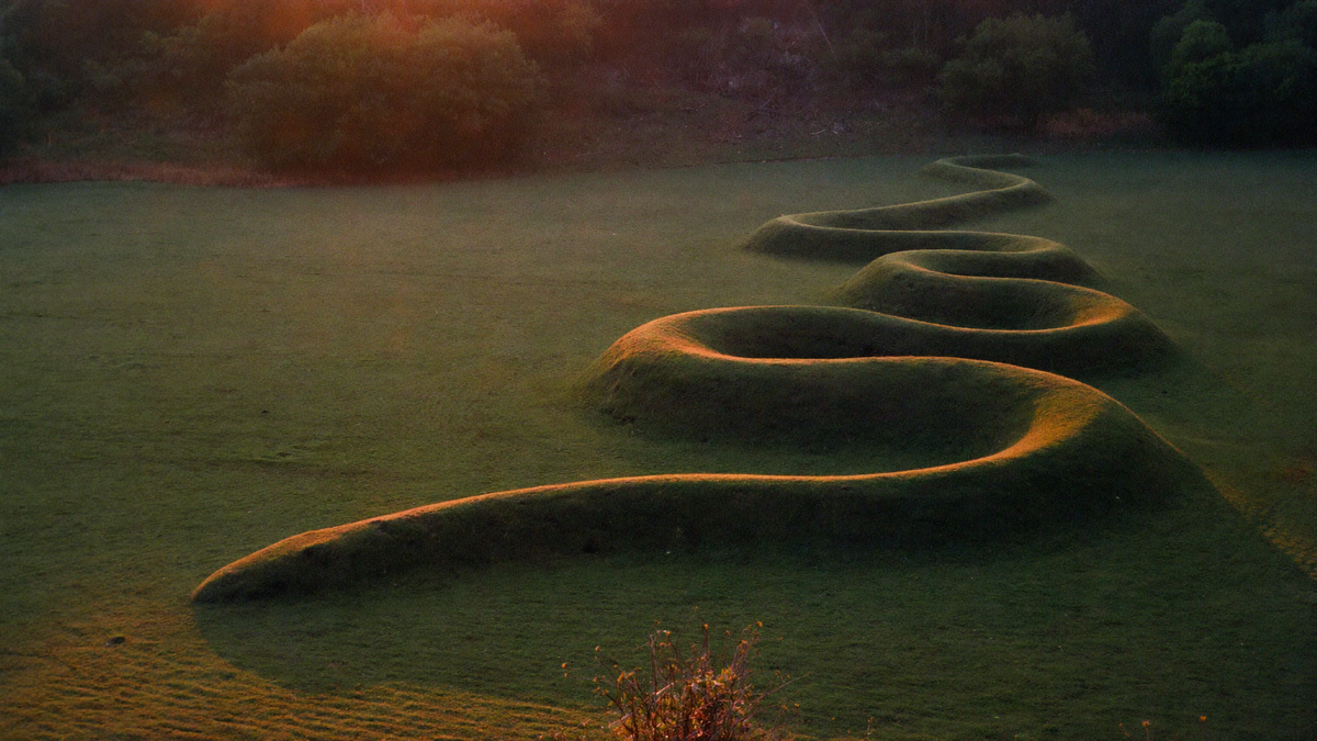 Grass-covered serpent-shaped earthwork winding across a meadow at dusk