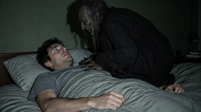 Young man lying awake and frozen in bed while a shadowy old hag leans over his chest