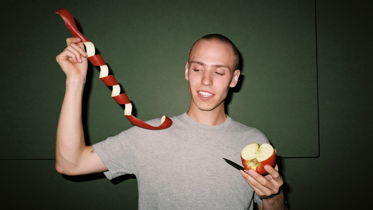 A young man holds up a long unbroken red apple peel spiral, a halved apple and paring knife in his other hand, against a dark green wall