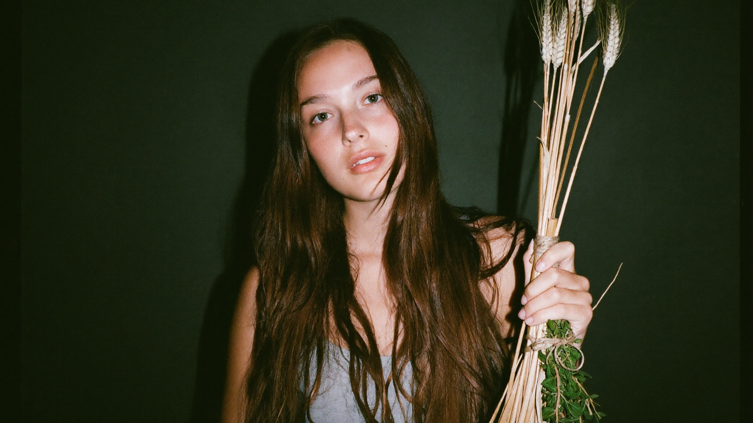 A young woman holding a tied bundle of dried wheat and herbs against a dark green wall