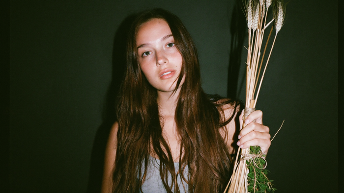 A young woman holding a tied bundle of dried wheat and herbs against a dark green wall