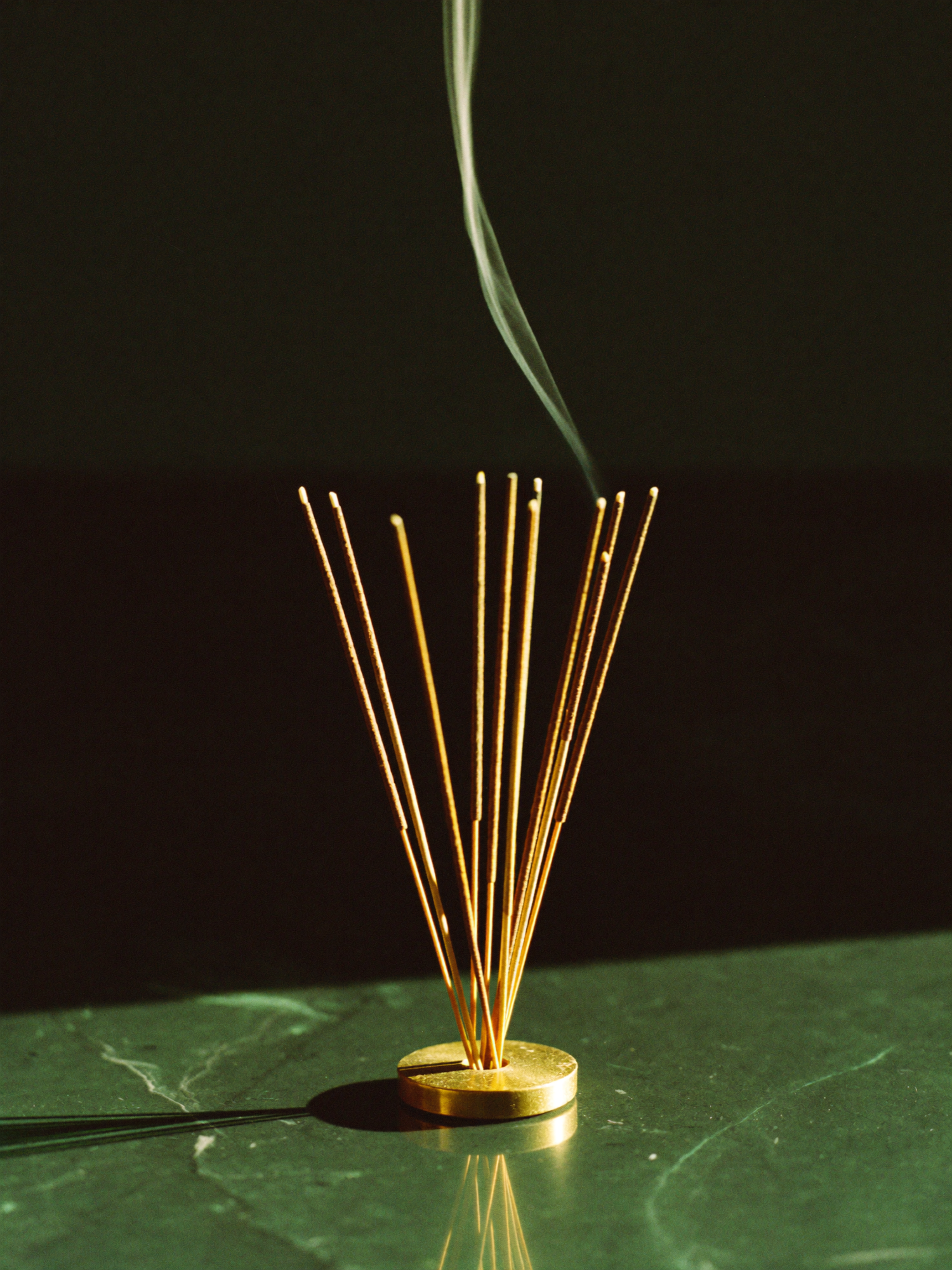 A stick of planetary incense smoldering on polished dark marble, a thin ribbon of smoke rising against a shadowed emerald-green background.