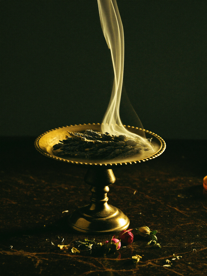 Smoldering white sage bundle in an antique brass dish with dried rose buds, lit from the side by candlelight