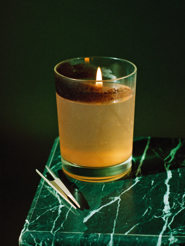 Seven-day glass ritual candle burning with soot on the upper glass, wick trimmers resting on dark marble beside it