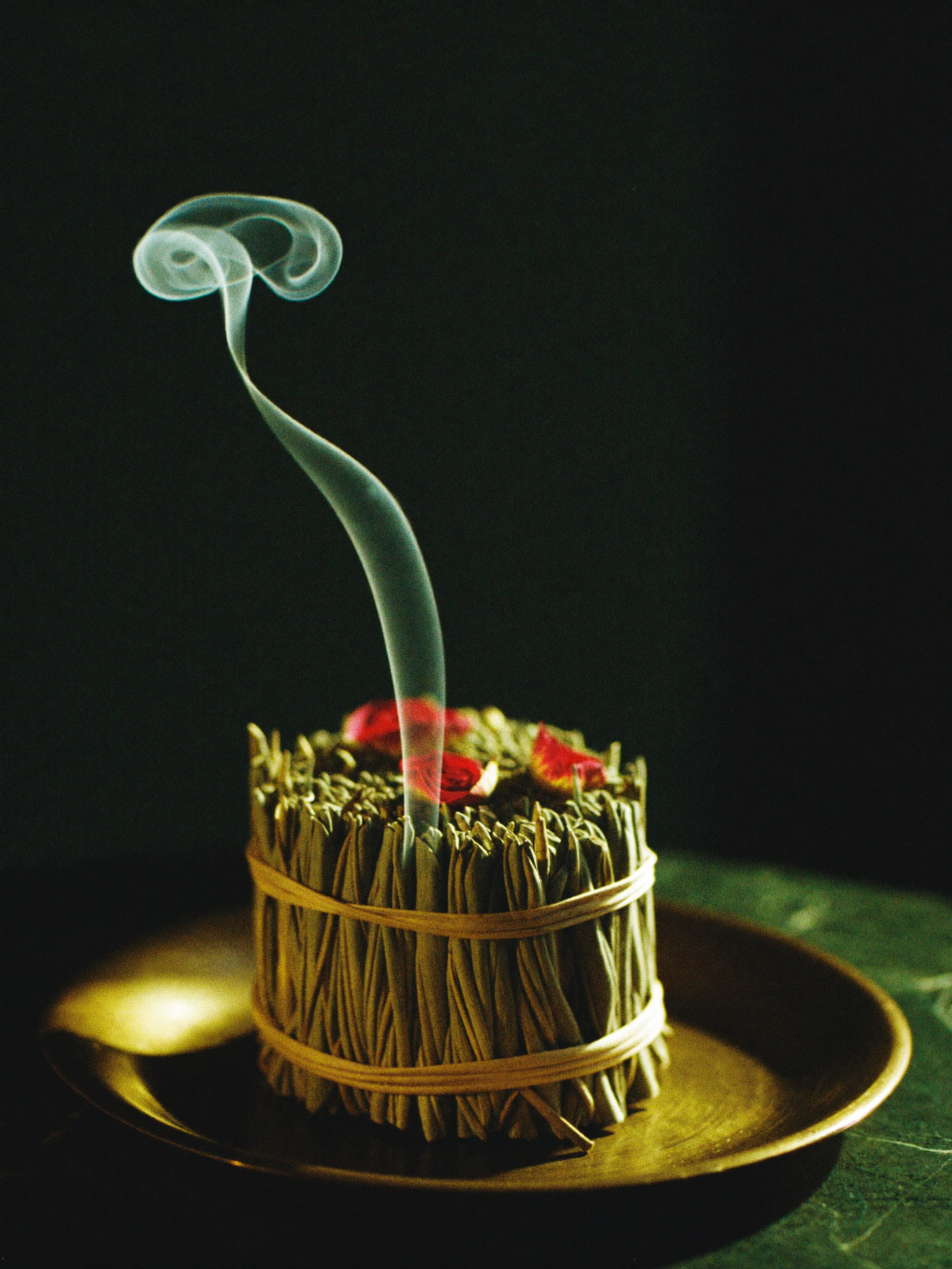 A smoldering smudge stick resting on polished dark marble, a curl of smoke lit by warm golden light against a deep emerald-green background.