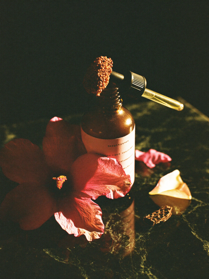 A small glass bottle of devotional oil on polished dark marble, lit by a single warm golden light against a deep emerald-green background falling into shadow.