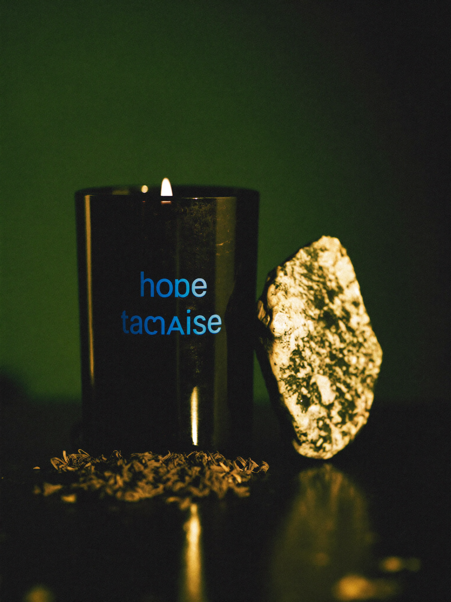 A banishing candle burning on polished dark marble, its flame lit by warm golden light against a shadowed emerald-green background.
