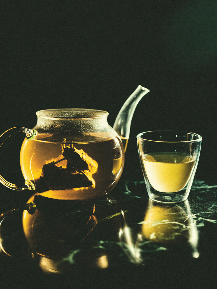 A clear glass teapot and cup on polished dark marble, warm golden light glowing through the tea against a deep emerald-green background.