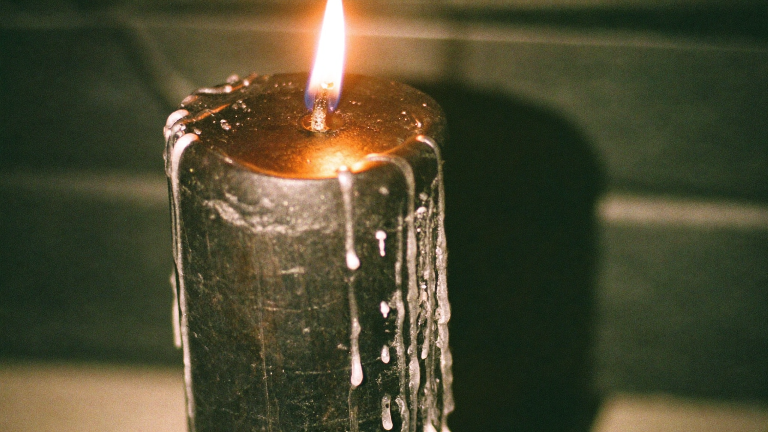 A black pillar candle burning close up with wax running down its side against a dark green wall