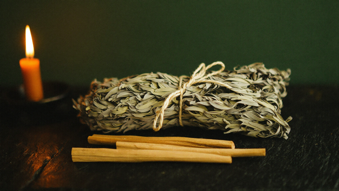 A tied white sage bundle and three palo santo sticks resting on dark stone beside a burning candle