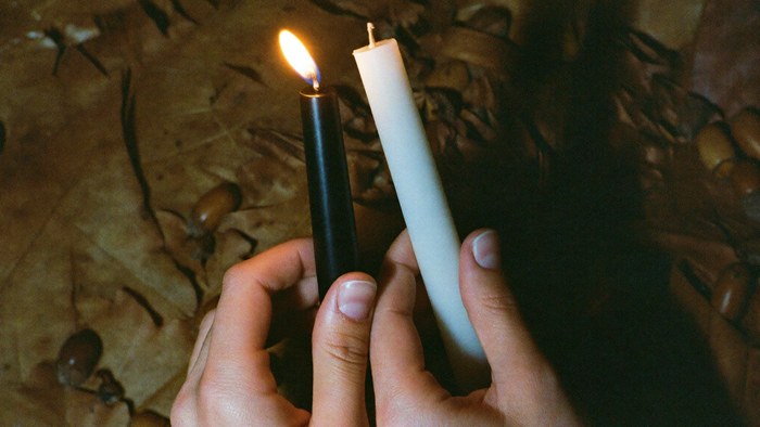 Hands holding a lit black taper candle and an unlit white taper candle above fallen oak leaves and acorns