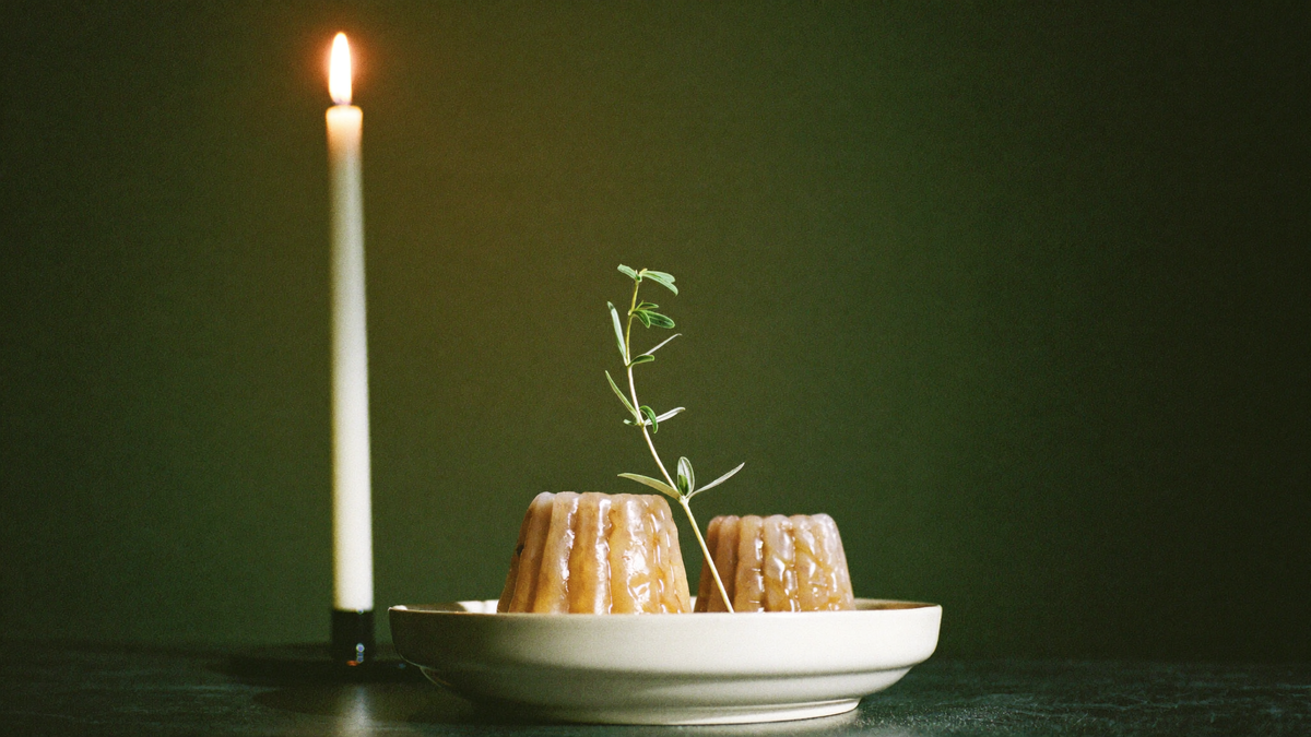 Honey cakes on a pale dish with an olive sprig beside a lit white candle against a dark green wall