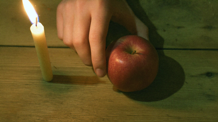 A hand setting a red apple down beside a burning candle stub on a worn wooden table