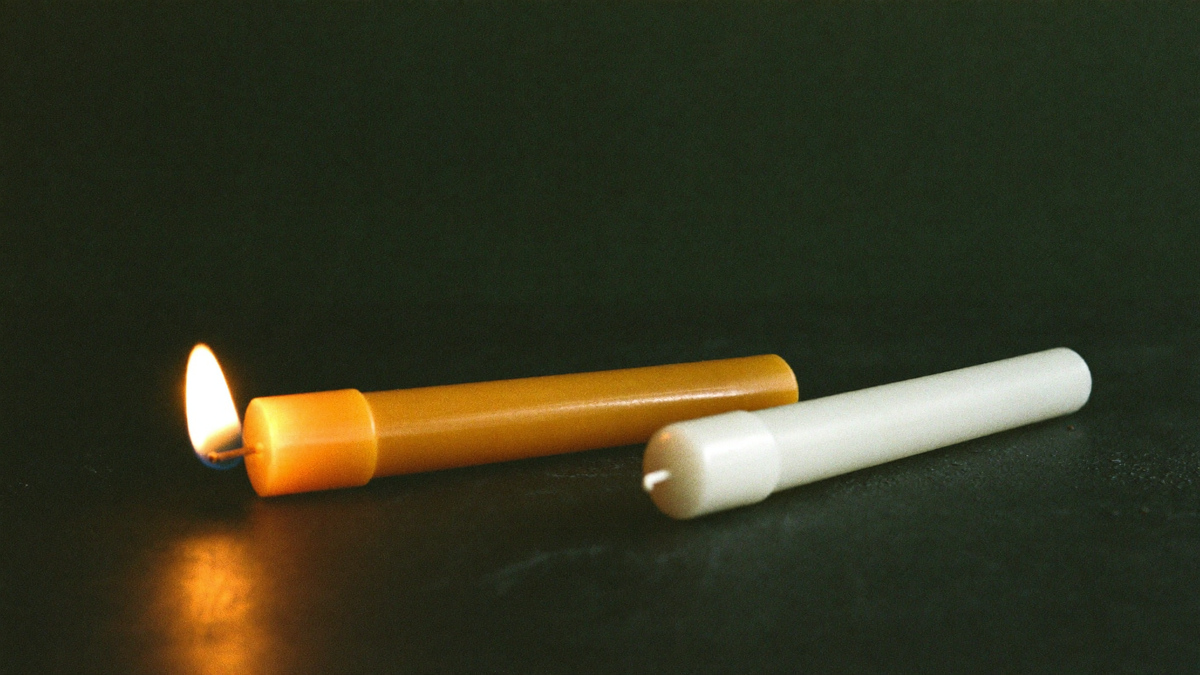 A golden beeswax taper candle burning beside an unlit white taper candle on dark stone
