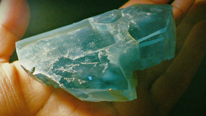 Rough blue-green apatite crystal with glassy fracture faces held in an open palm in warm light