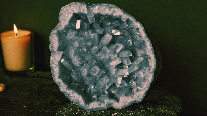 Pale blue celestite geode with glassy blocky crystals resting on dark stone beside a lit candle