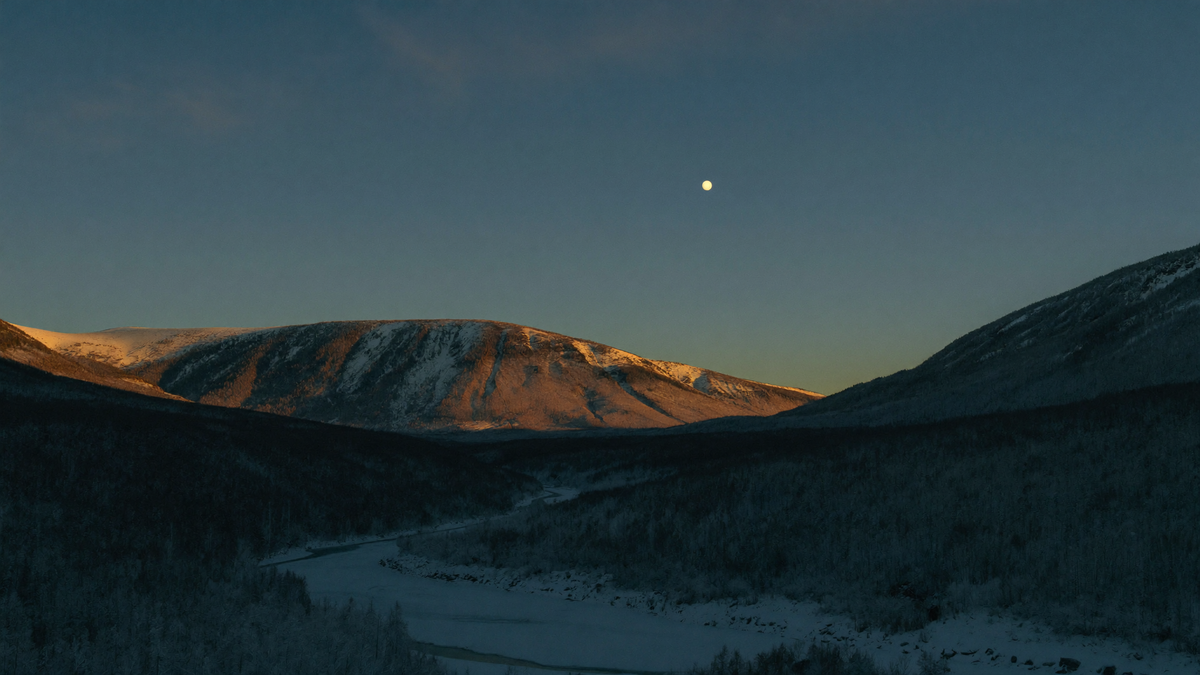 Snow-covered Norwegian valley at blue hour with a frozen river below and a single pale light above the far ridge