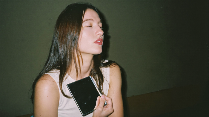 Woman with closed eyes holding a small hand mirror against her chest in front of a dark green wall