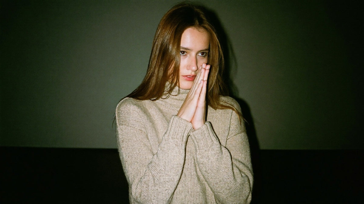 Young woman in an oatmeal knit sweater pressing her palms together in front of her lips against a dark green wall