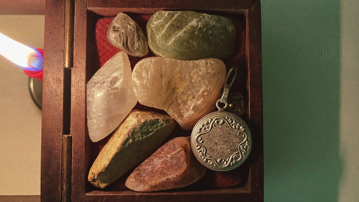 Old tumbled stones, a quartz point and a tarnished silver locket in an open wooden box lit by candlelight