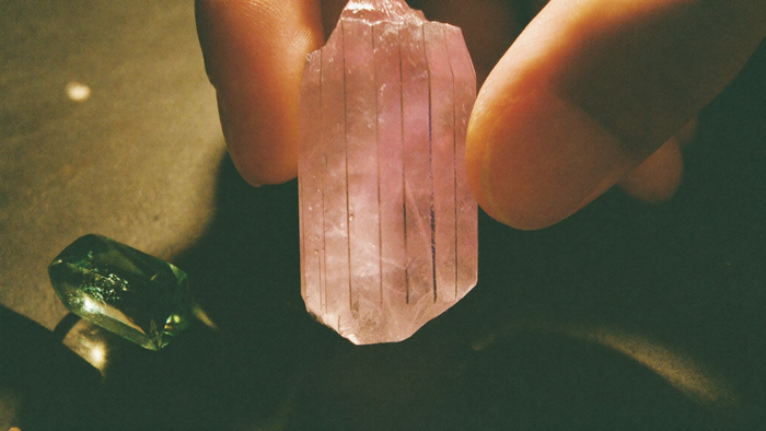 Fingertips holding a pale pink kunzite crystal with fine lengthwise striations up to the light, a green crystal beside it
