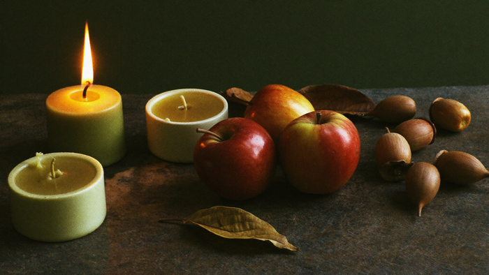 Beeswax tea light candles, apples, acorns and dried oak leaves on a dark stone surface lit by one candle
