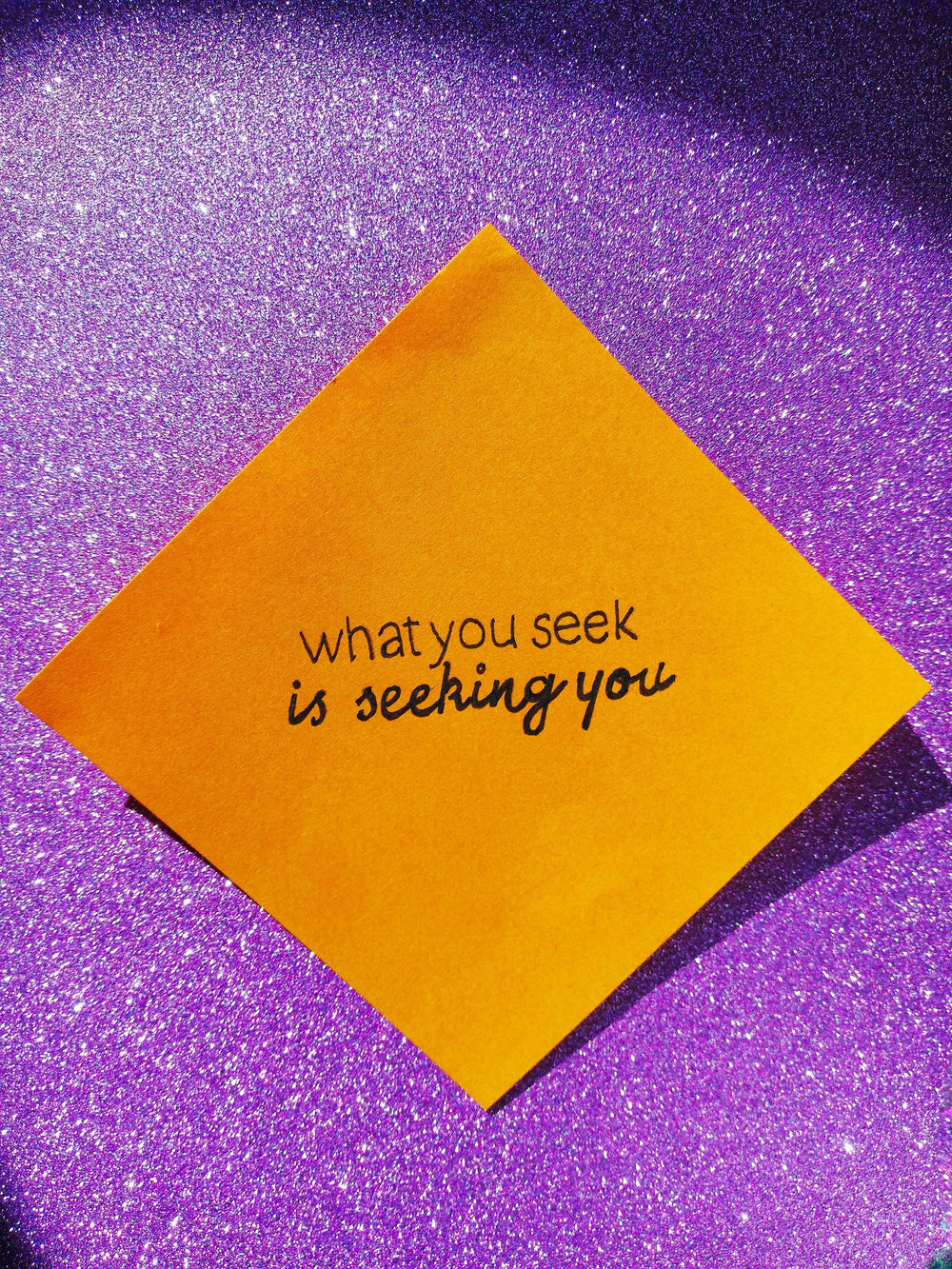 Orange sticky note reading what you seek is seeking you on a sparkling purple glitter background