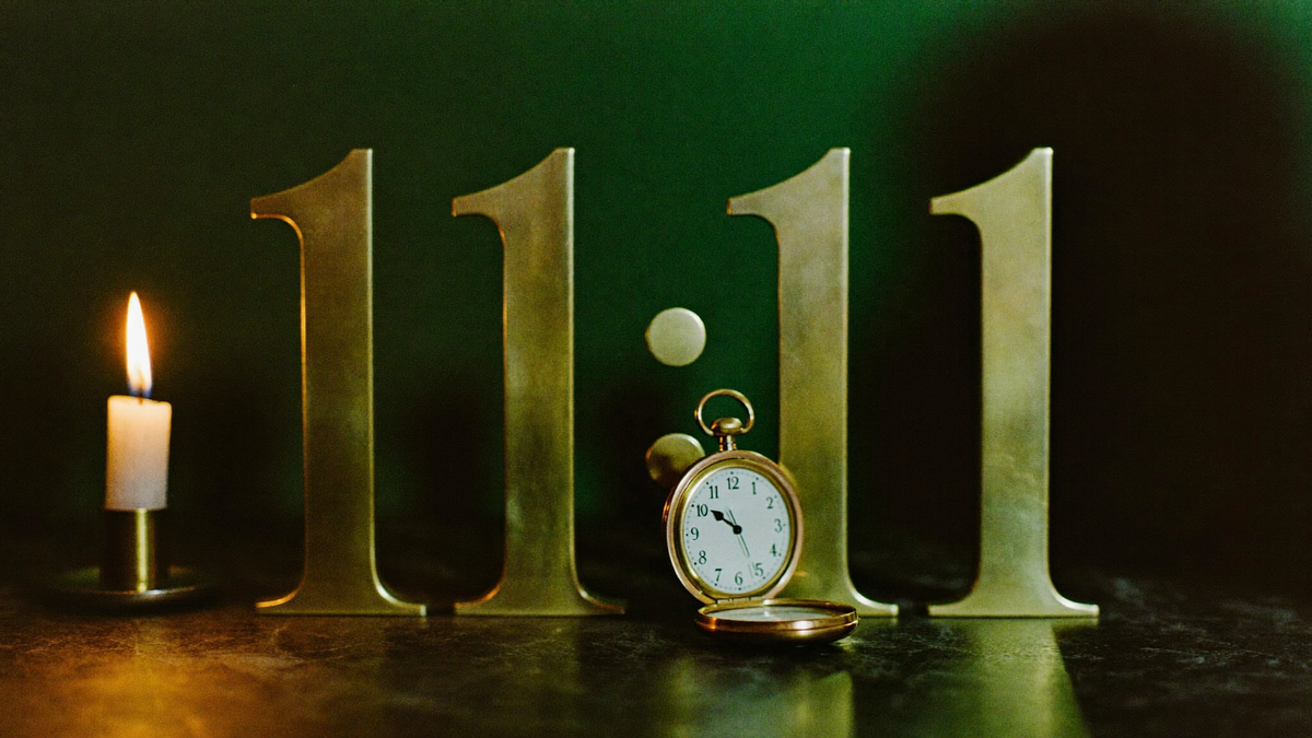 Large brass numerals reading 11:11 against a dark green wall with an open pocket watch and a lit candle in front
